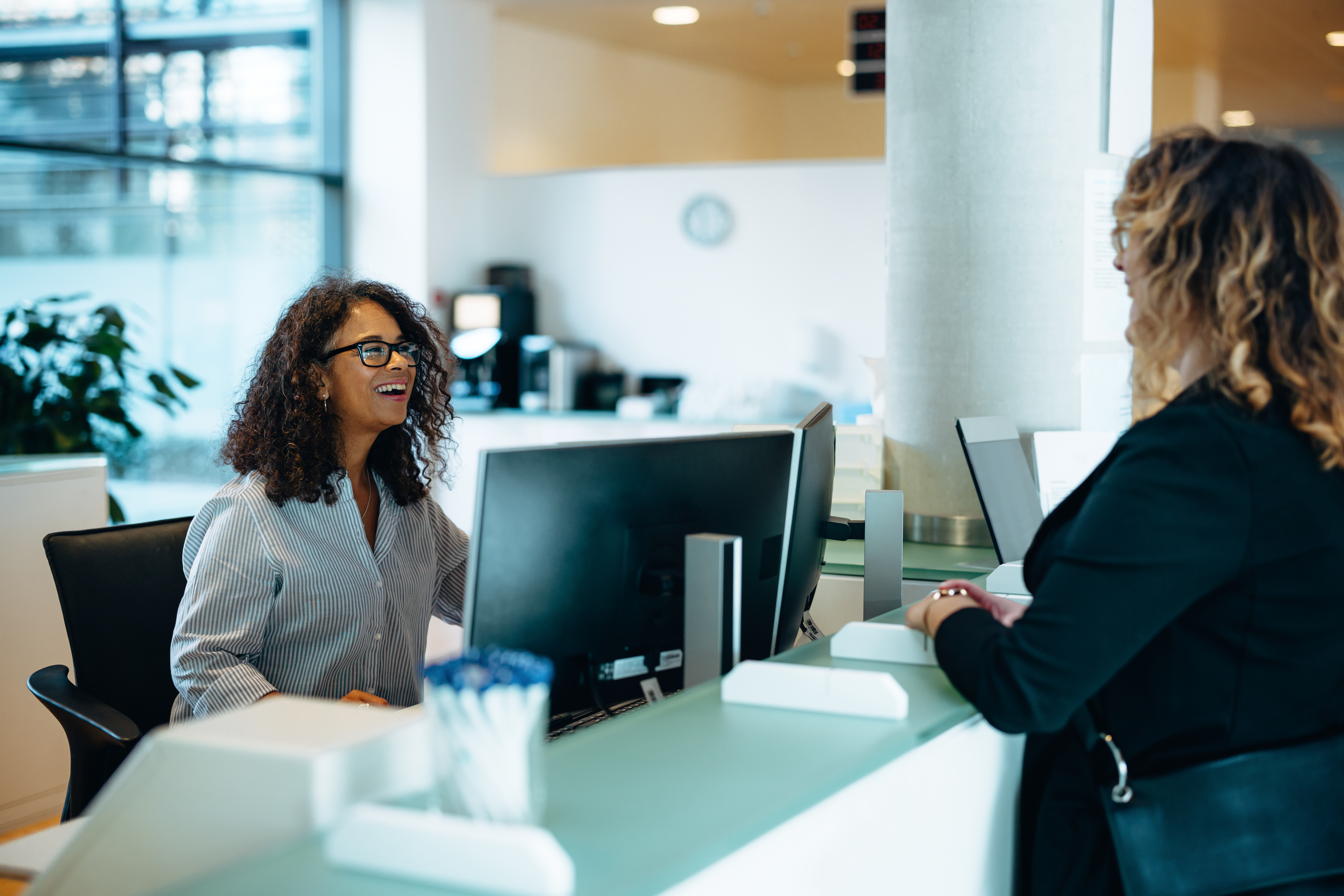 Friendly administrator assisting woman at reception desk Receptionist assisting a woman standing at front desk. Woman standing at the reception talking with a friendly receptionist behind the desk of municipality office.